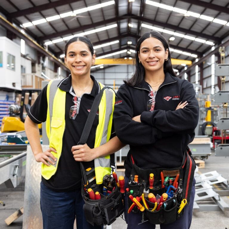Two female electrical apprentices in a workshop, wearing safety gear and tool belts, smiling confidently.