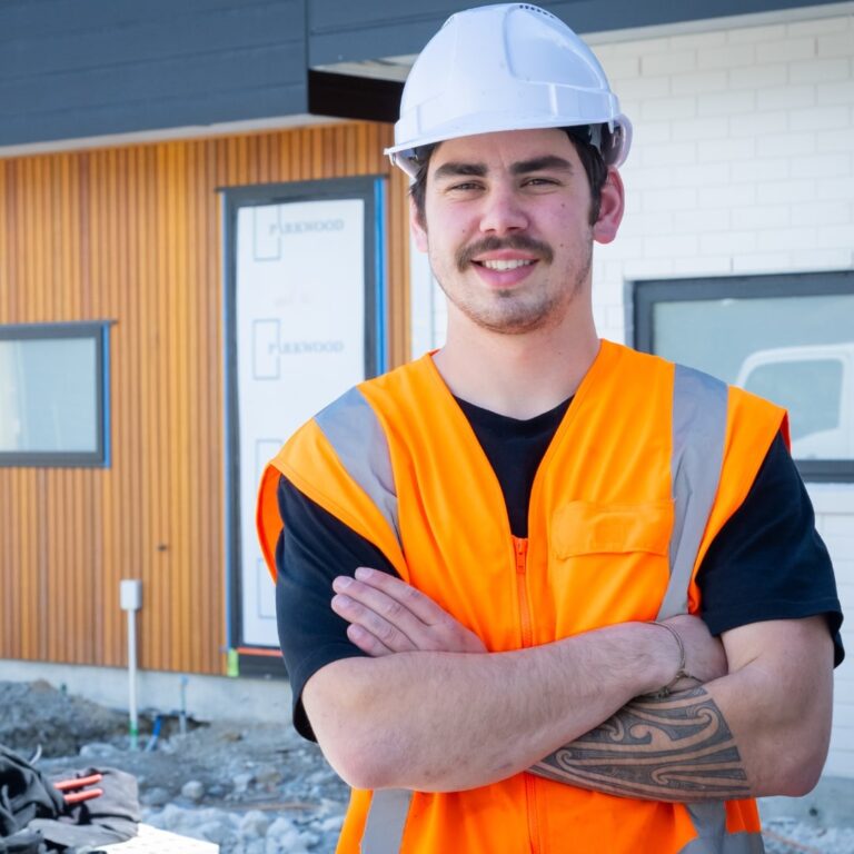 Plumbing apprentice in hi-vis vest and hard hat standing confidently with arms crossed at a building site.