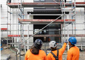 Three EarnLearn trainees in orange high-vis and helmets looking at scaffolding and a partially built house, with one trainee pointing toward the structure.
