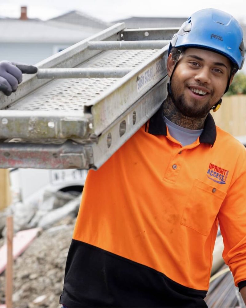 Smiling scaffolder in hi-vis and helmet carrying scaffolding equipment on a construction site.