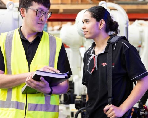Two apprentices in hi-vis vests discussing notes inside a workshop.
