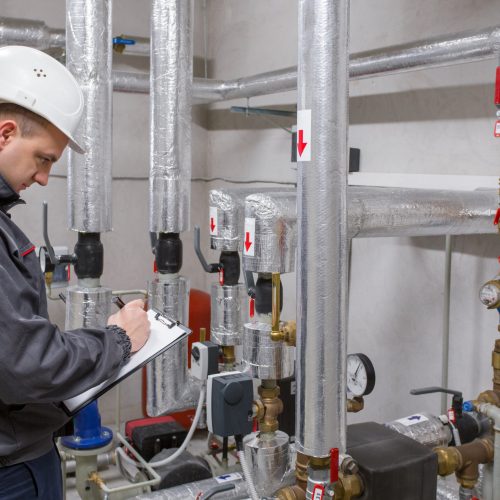 Engineer in a hard hat inspecting industrial piping systems and taking notes on a clipboard.